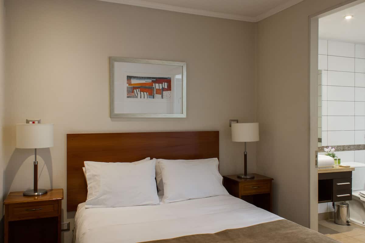 Habitación de hotel con cama, dos mesitas de noche con lámparas, una obra de arte enmarcada en la pared y un baño visible a través de una puerta abierta.