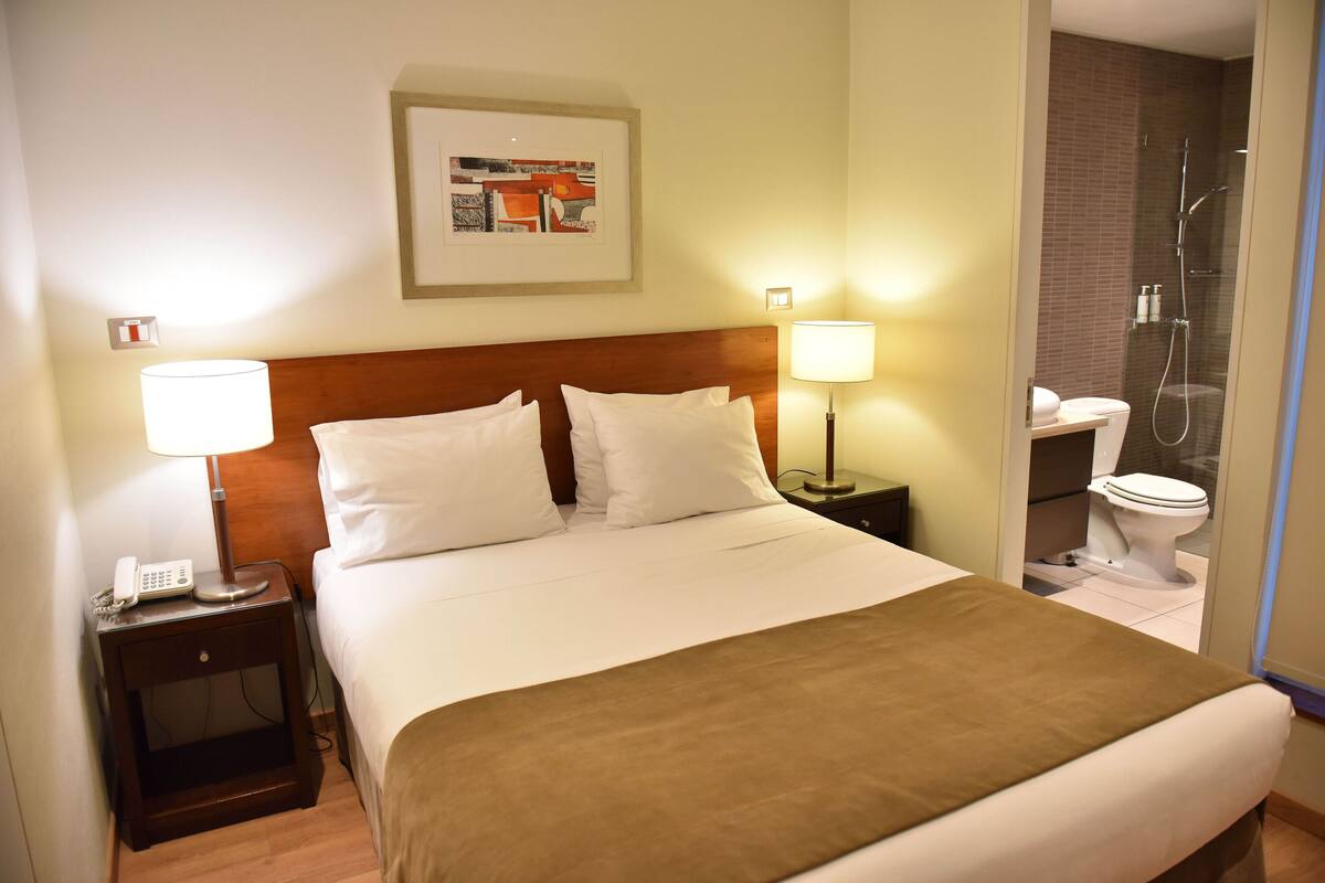 Habitación de hotel con cama, dos lámparas de noche, mesita de noche con teléfono, cuadro en la pared y baño visible a través de una puerta abierta.