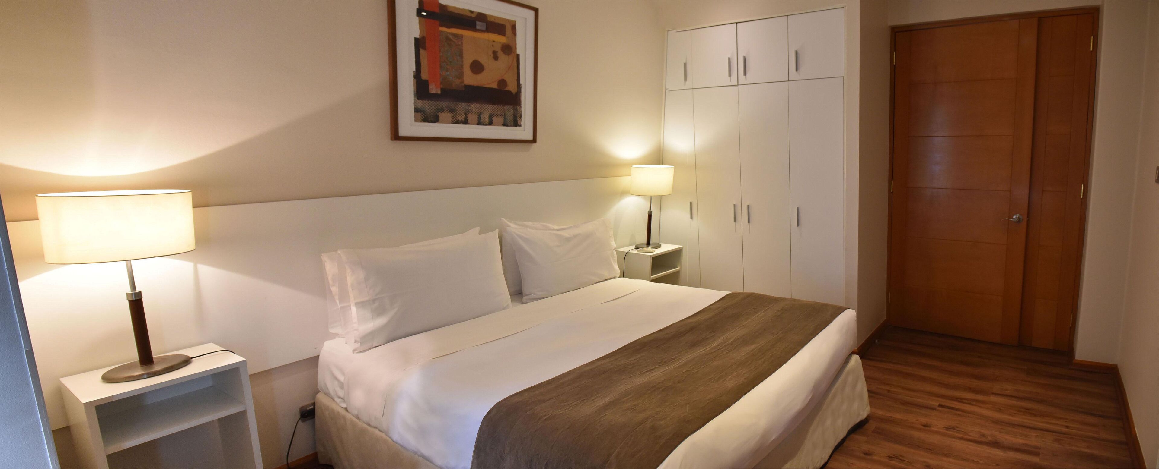 Een net opgemaakt bed met witte lakens en een bruine plaid, twee bedlampjes, een houten deur en een witte garderobe.