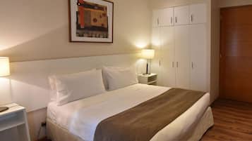 Een net opgemaakt bed met witte lakens en een bruine plaid, twee bedlampjes, een houten deur en een witte garderobe.