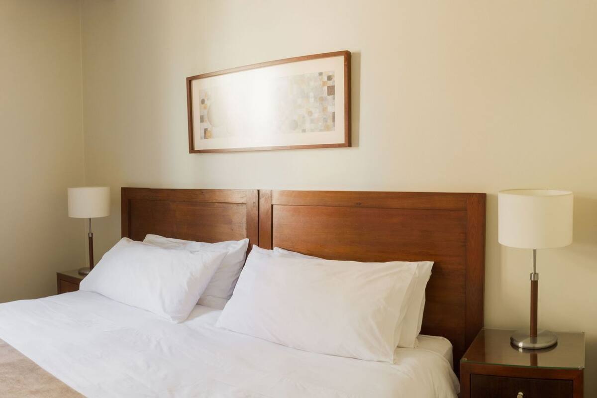 Habitación de hotel con cama, dos mesitas de noche con lámparas, una ventana con cortinas, un radiador y un cuadro en la pared.