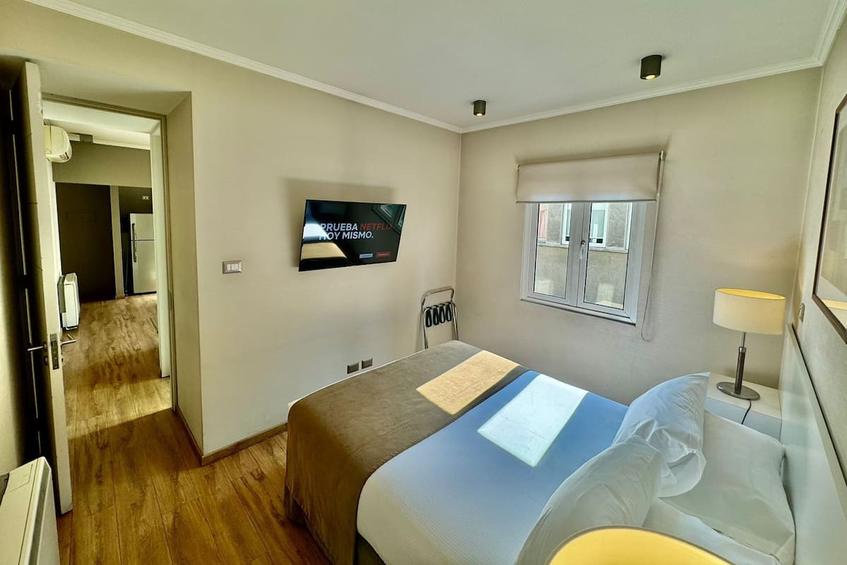 Habitación de hotel con cama, televisor montado en la pared, ventana con vista y puerta que da a otra habitación.