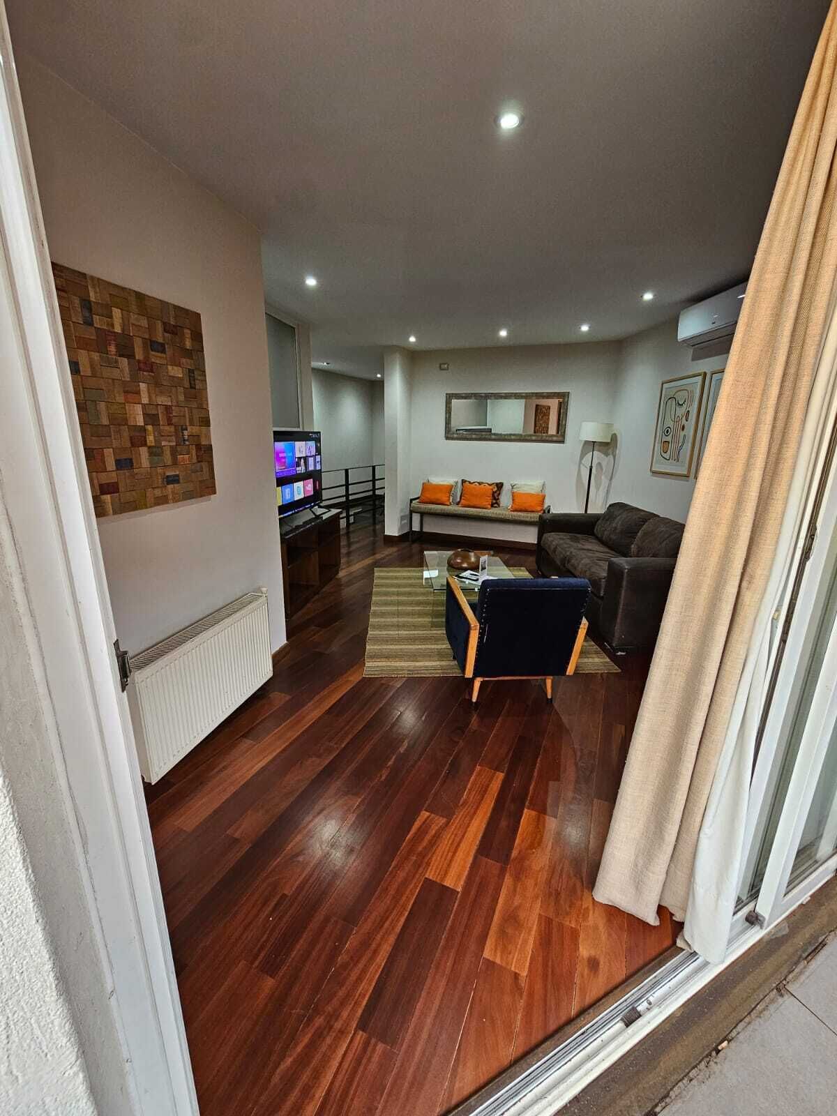 Uma sala de estar moderna com piso de madeira, um sofá, uma televisão e uma área de jantar ao fundo.