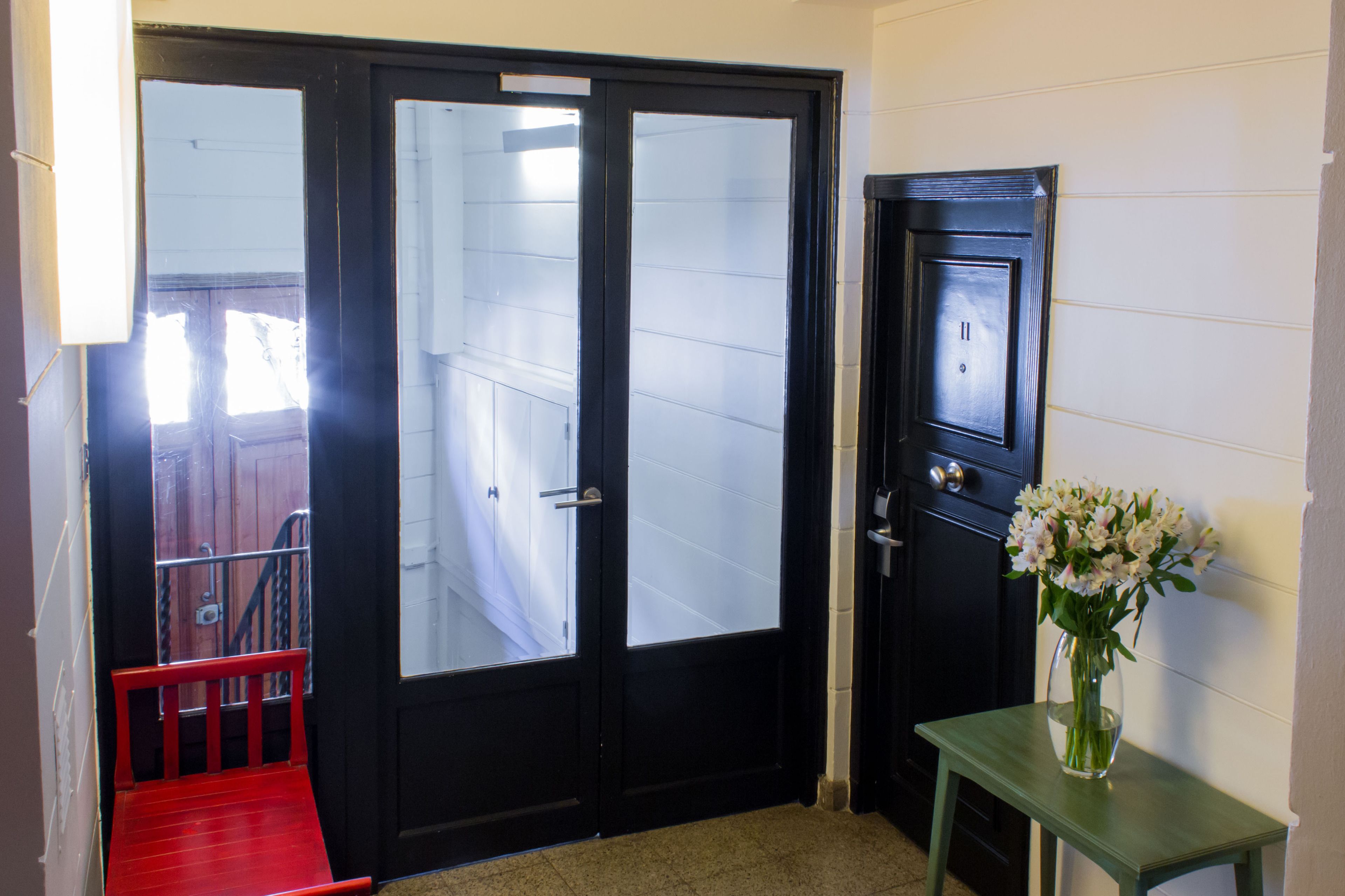 A black door with frosted glass panels, a small green table with a vase of flowers, and a red bench outside.