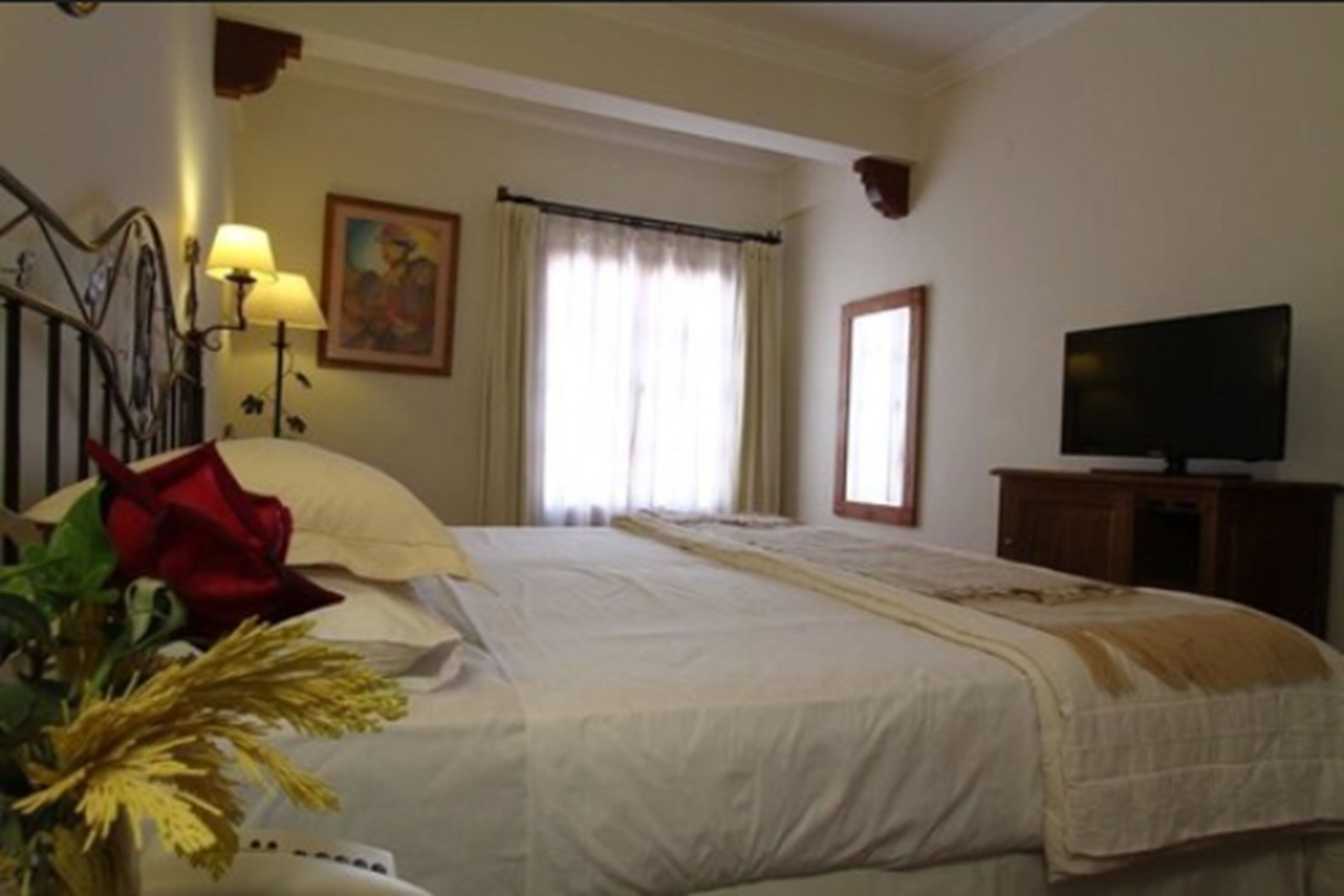 Une chambre d’hôtel avec un lit, une télévision, une lampe, un tableau et une fenêtre avec des rideaux.