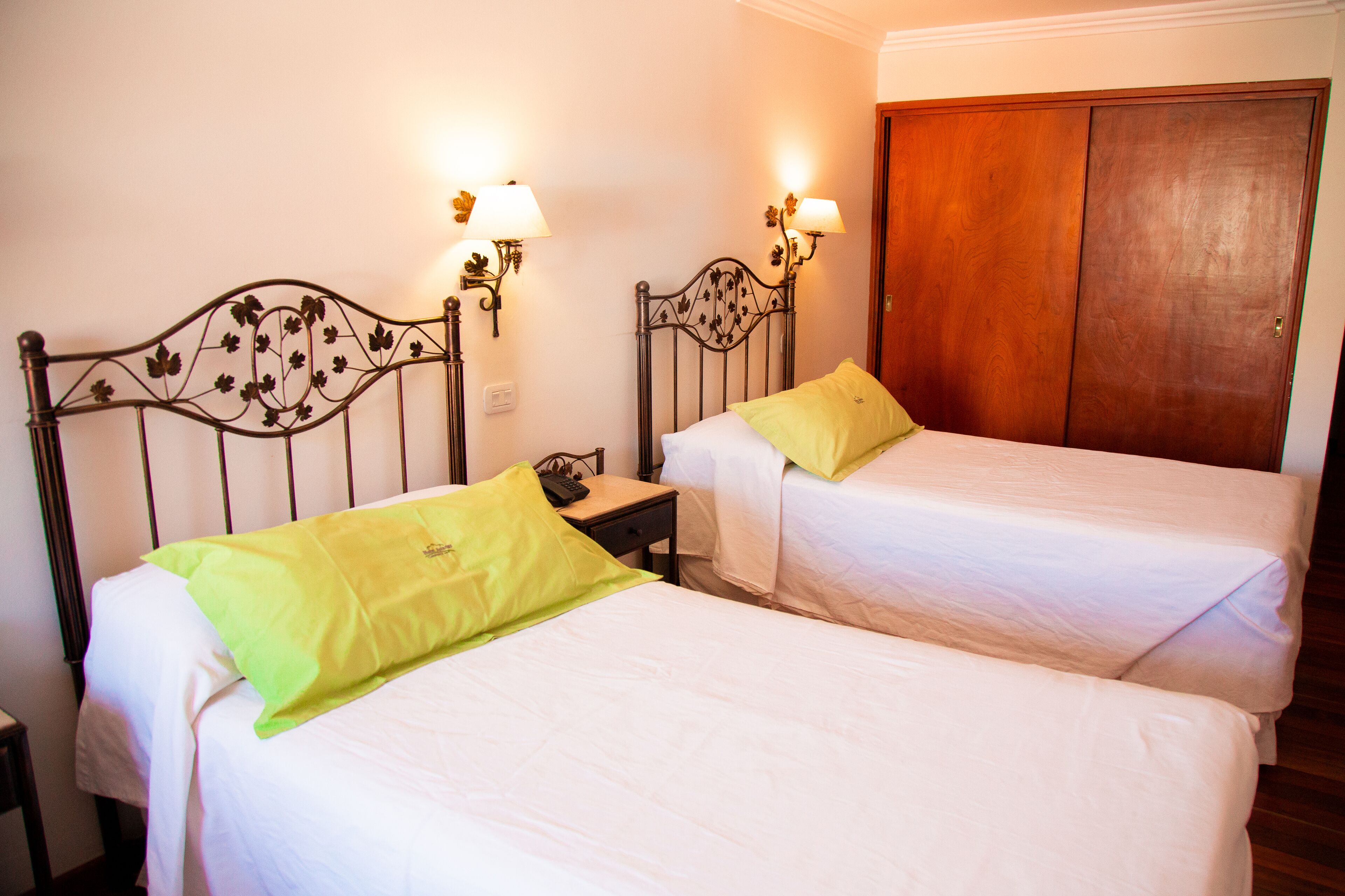 A hotel room with two single beds, each with a pillow and a bedside table. There are wall-mounted lamps on either side of the beds.