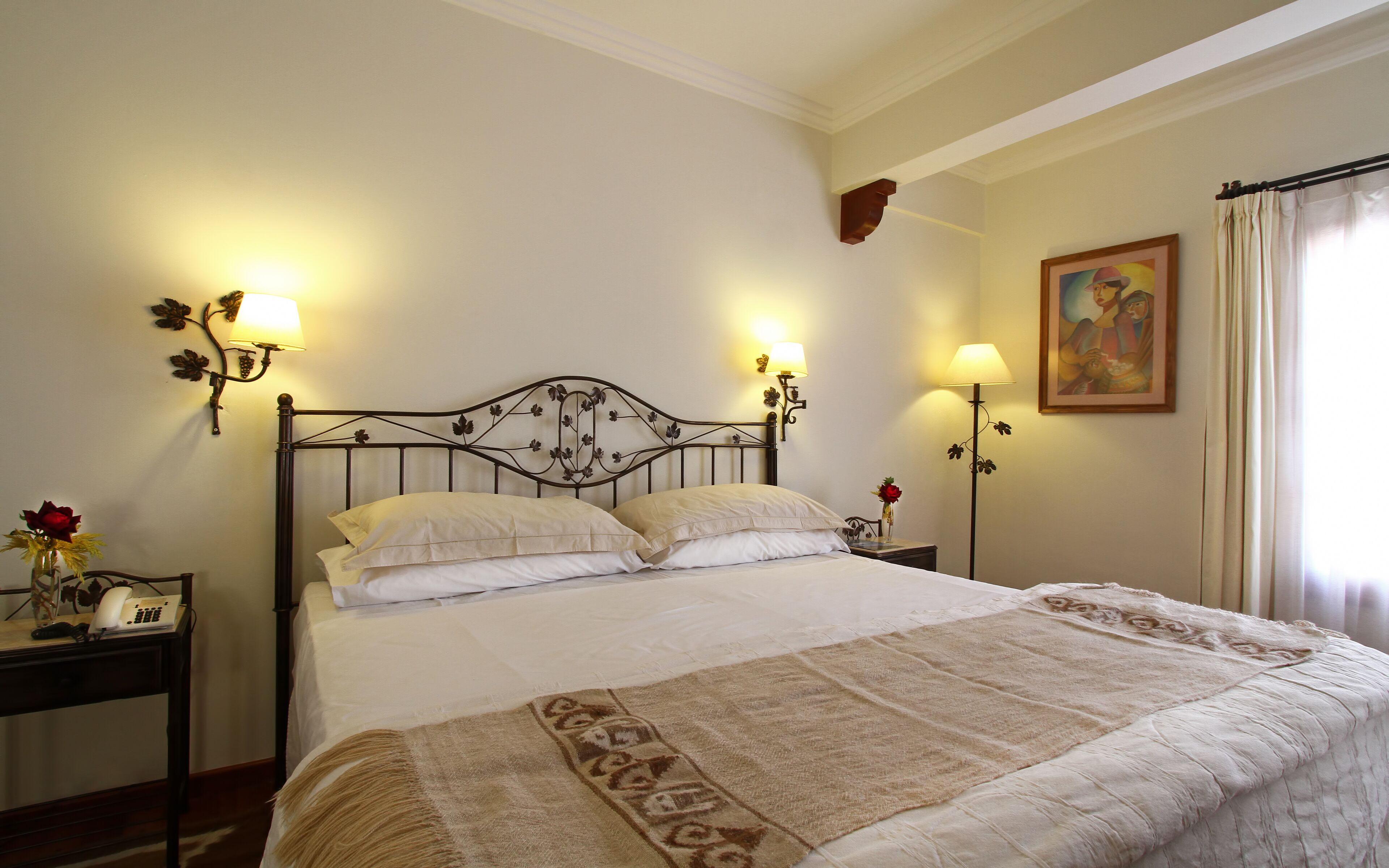 A neatly made bed with a patterned bedspread, two bedside lamps with floral designs, a nightstand with a telephone, and a painting on the wall.