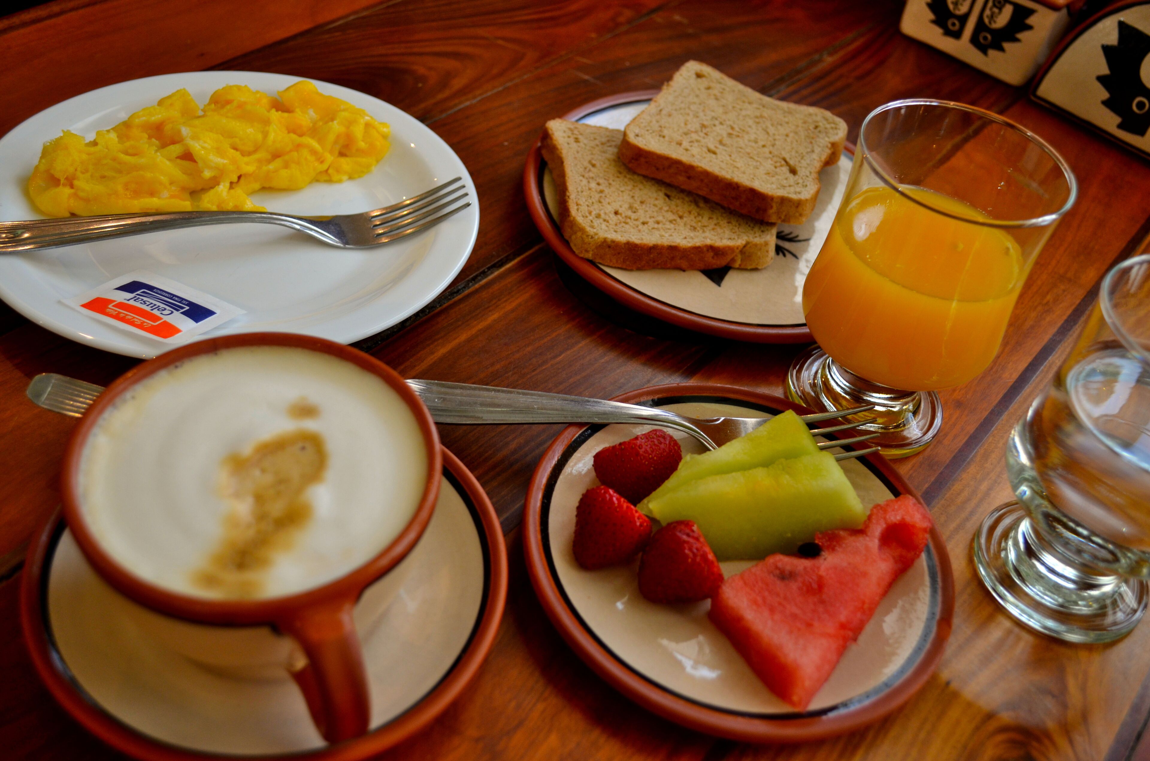 Un desayuno con huevos revueltos, tostadas, un vaso de jugo de naranja, una taza de café y un plato con fresas y sandía.