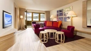 Une chambre d’hôtel moderne avec un canapé violet, deux tabourets ronds en bois, un téléviseur à écran plat et une grande fenêtre avec des rideaux.
