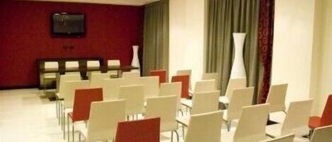 A conference room with rows of white and red chairs, a flat-screen TV on the wall, and a large window with curtains.