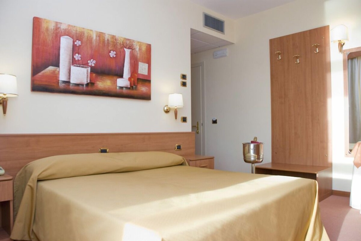 Una habitación de hotel con cama, un cuadro en la pared, una mesita de noche, una lámpara y un armario.