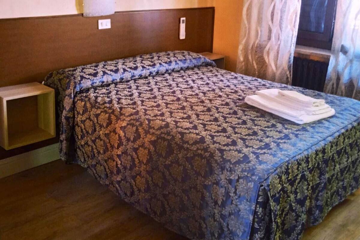 A hotel room with a wooden bed, a nightstand, a painting on the wall, and a window with curtains.