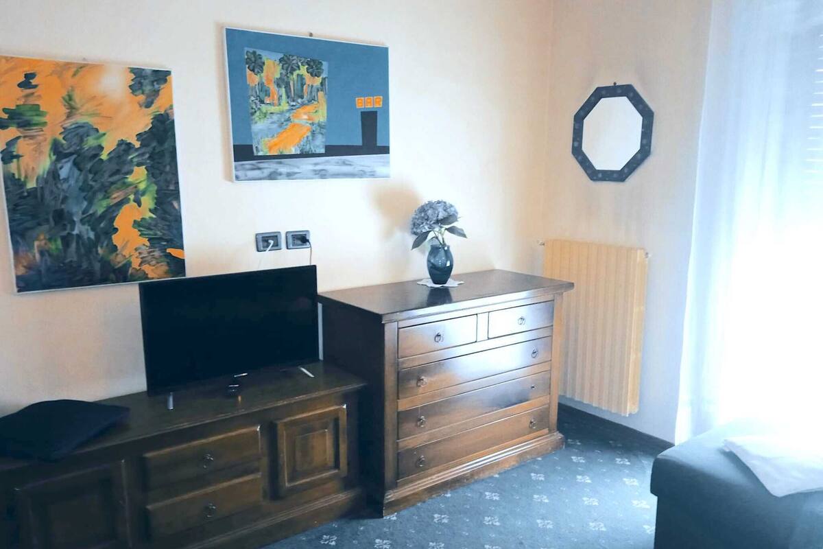 A hotel room with a wooden dresser, a flat-screen TV, a painting, a vase with a flower, and a radiator.