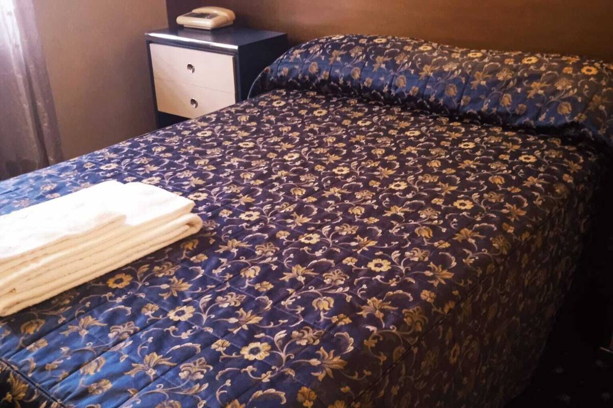A neatly made bed with a floral-patterned bedspread, a bedside table with a lamp, and a wall-mounted switch.