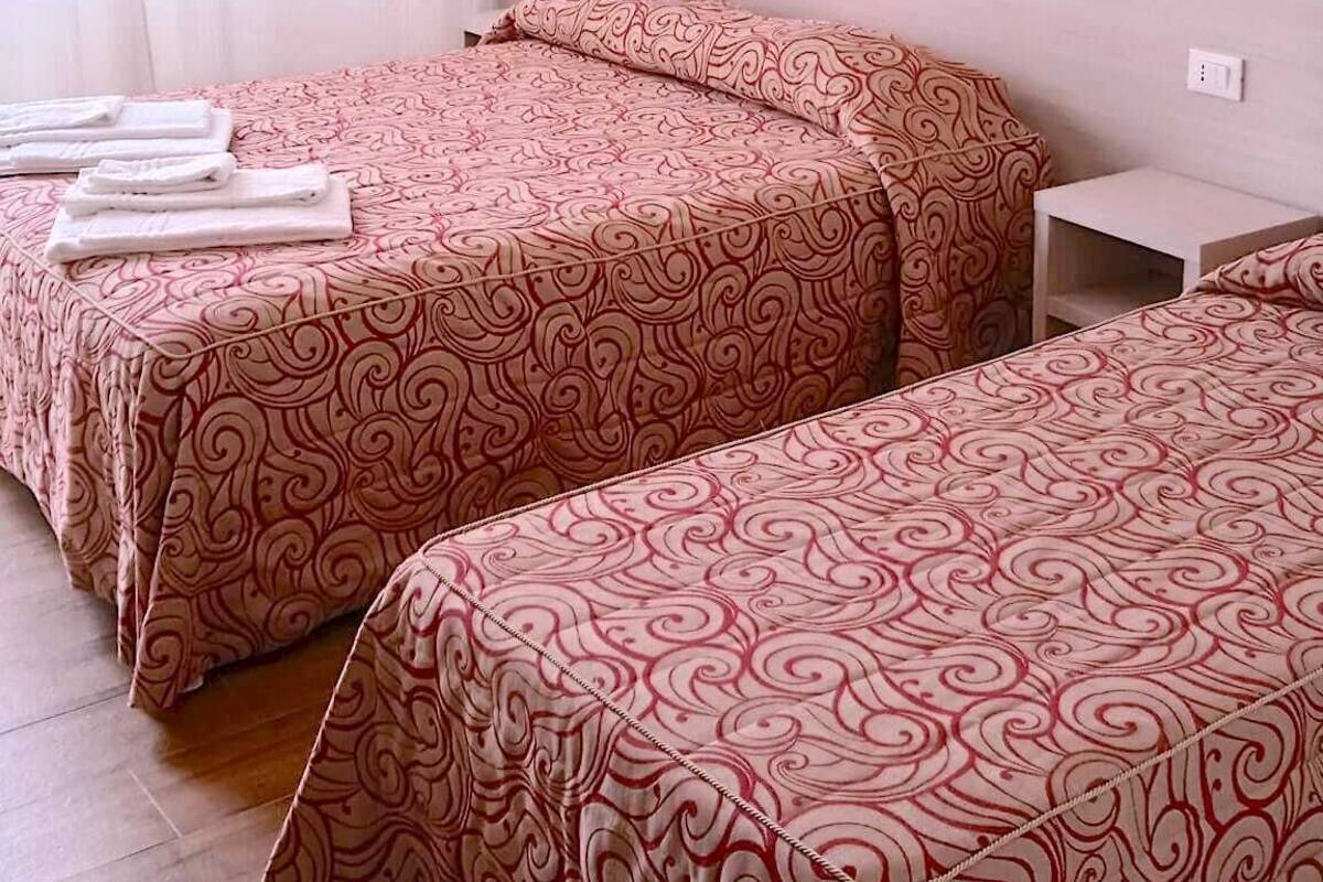 A hotel room with two beds, each with a patterned bedspread, a bedside table, a wall-mounted lamp, and a painting on the wall.