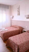 Una camera d'albergo con due letti, ciascuno con una coperta a motivi decorativi, un comodino, una lampada a parete e un quadro appeso.