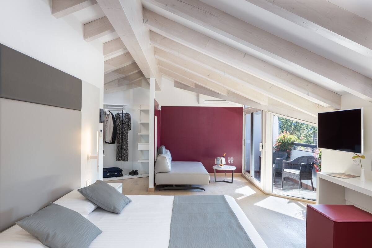 A modern hotel room with a bed, a seating area with a red wall, and a balcony with outdoor furniture.