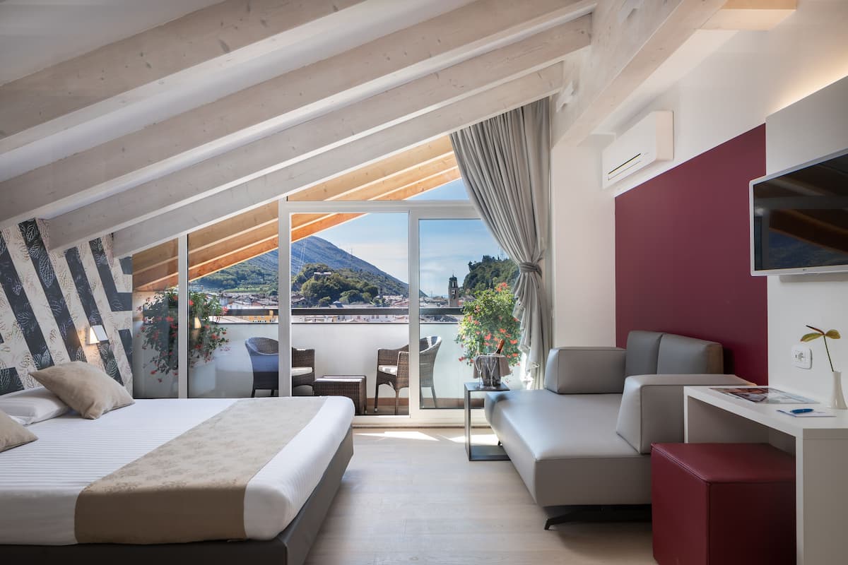 A modern hotel room with a large bed, a sofa, a desk, and a balcony with a mountain view.