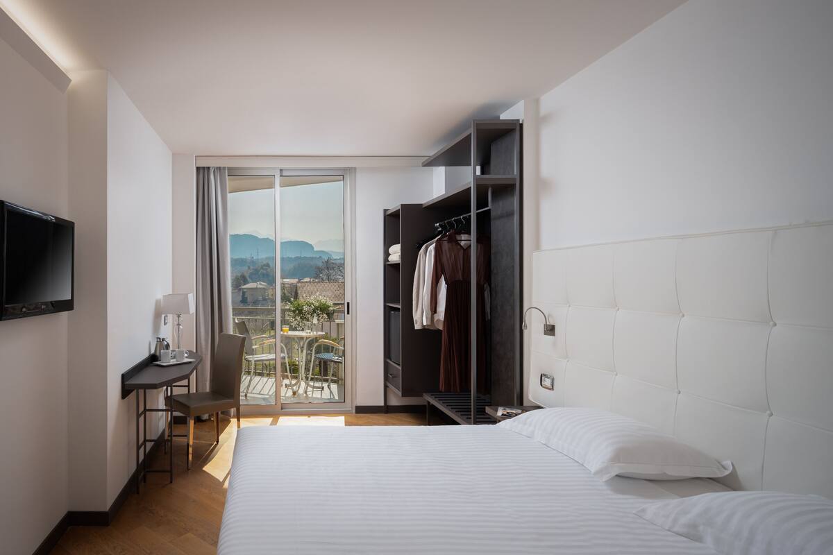 A hotel room with a bed, a desk, a chair, a TV, a wardrobe, and a balcony with a view of the mountains.