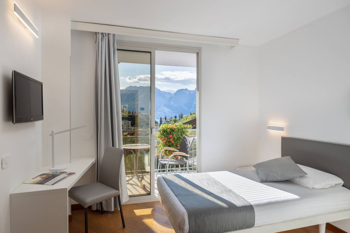 A bedroom with a bed, a desk, a chair, and a TV. There is a balcony with a table and chairs, and a view of mountains.