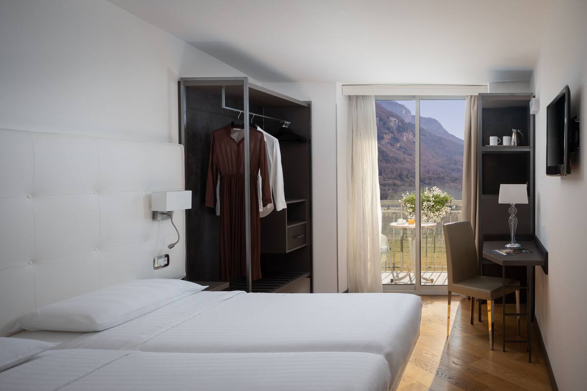 A hotel room with a bed, a desk, a chair, a wardrobe, and a balcony with a mountain view.