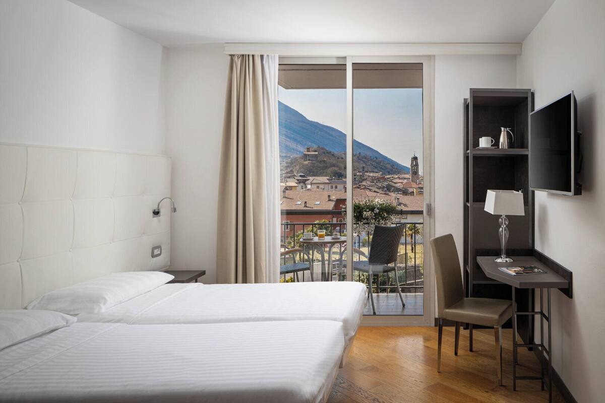 A hotel room with two beds, a desk, a chair, and a TV. The room has a large window with a view of a town and mountains.