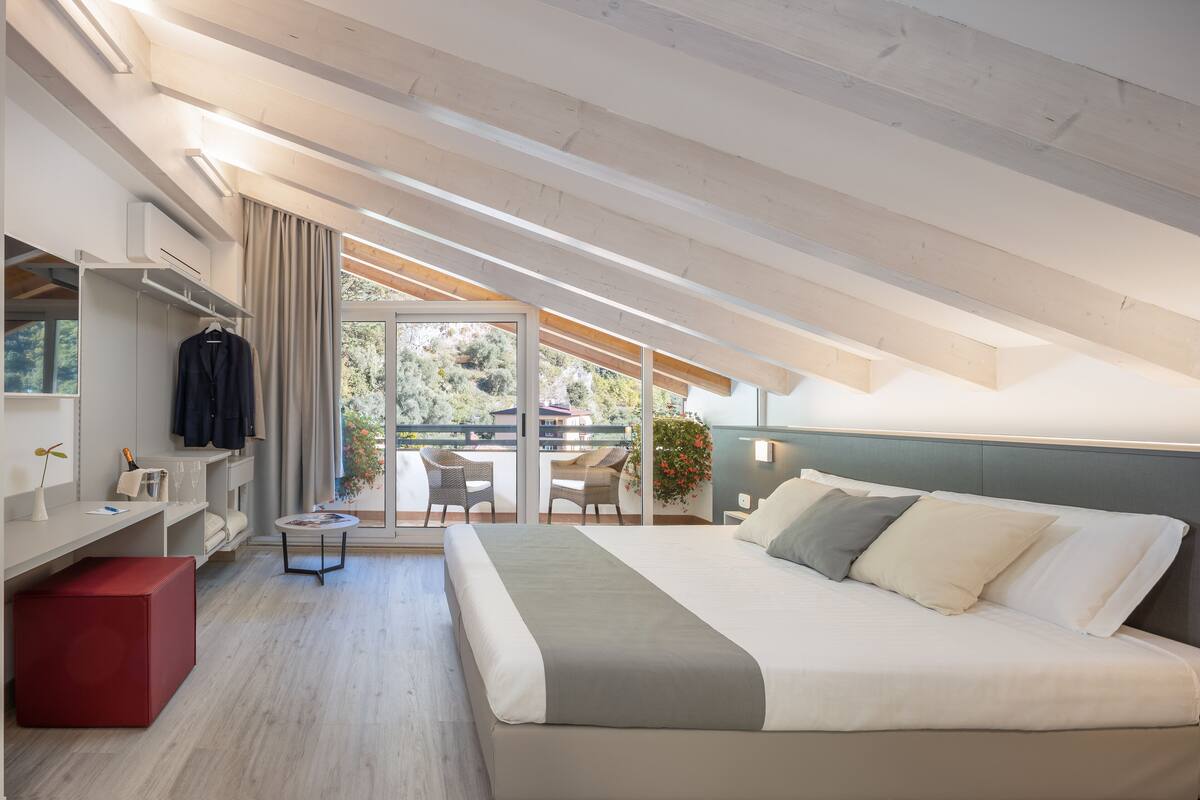 A modern bedroom with a large bed, a red ottoman, a small table, and a balcony with chairs.