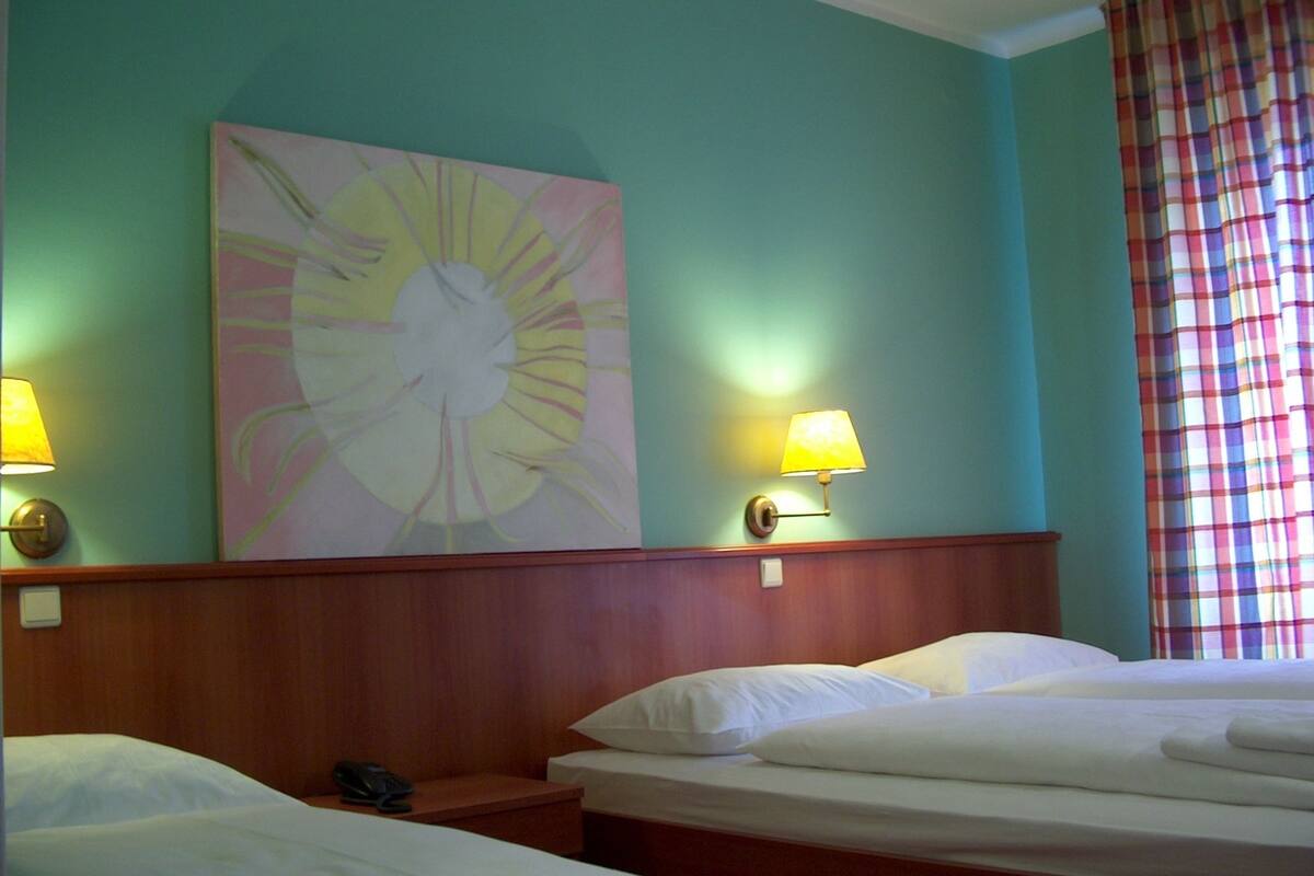 Quarto de hotel com duas camas, um quadro abstrato grande na parede e uma janela com cortina xadrez.