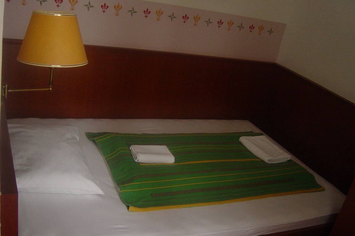 Uma cama de solteiro com um cobertor verde listrado e duas toalhas brancas dobradas. Um abajur amarelo está sobre uma mesa de cabeceira.