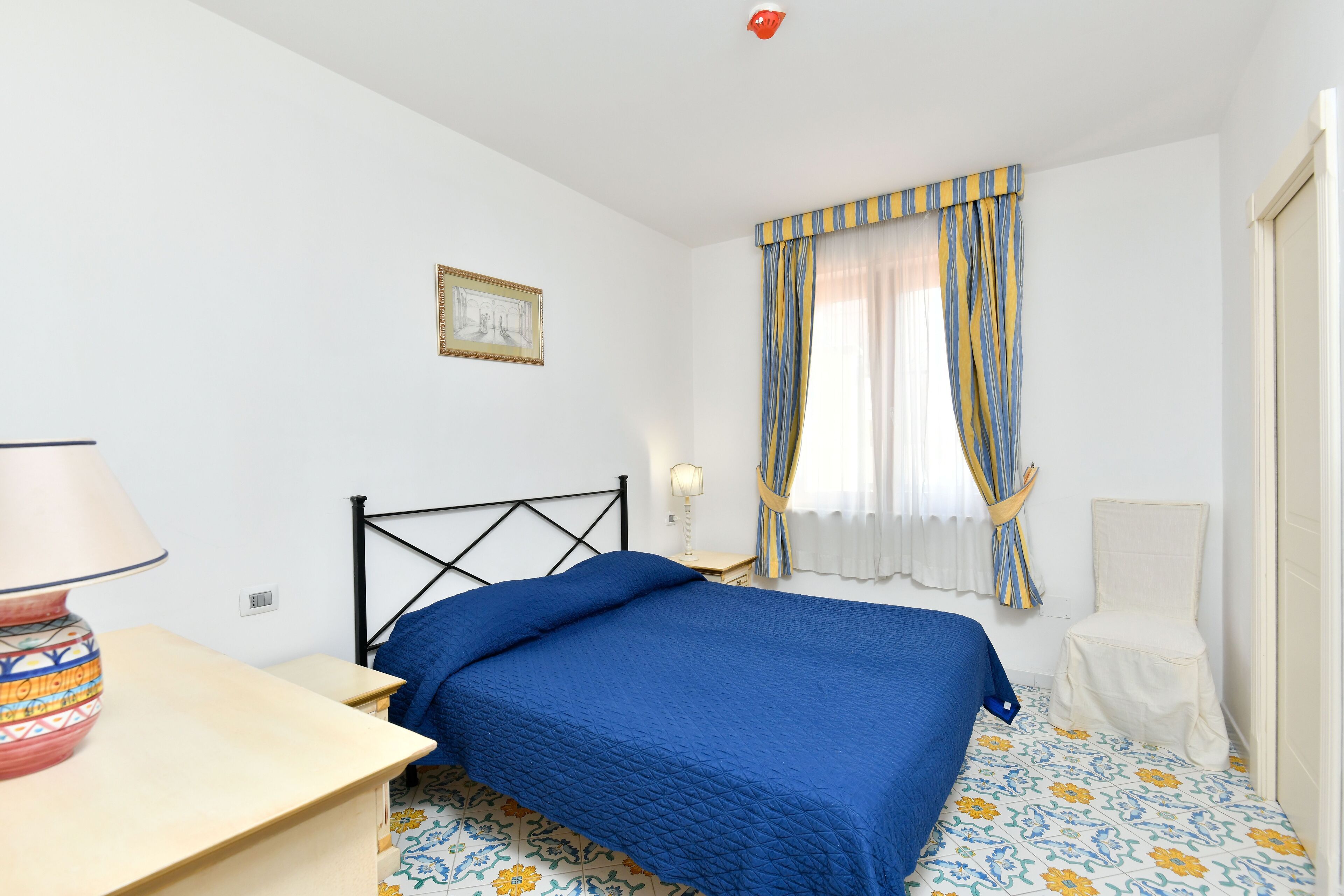 A bedroom with a bed, a bedside table, a lamp, a chair, a window with curtains, and a wall-mounted picture.