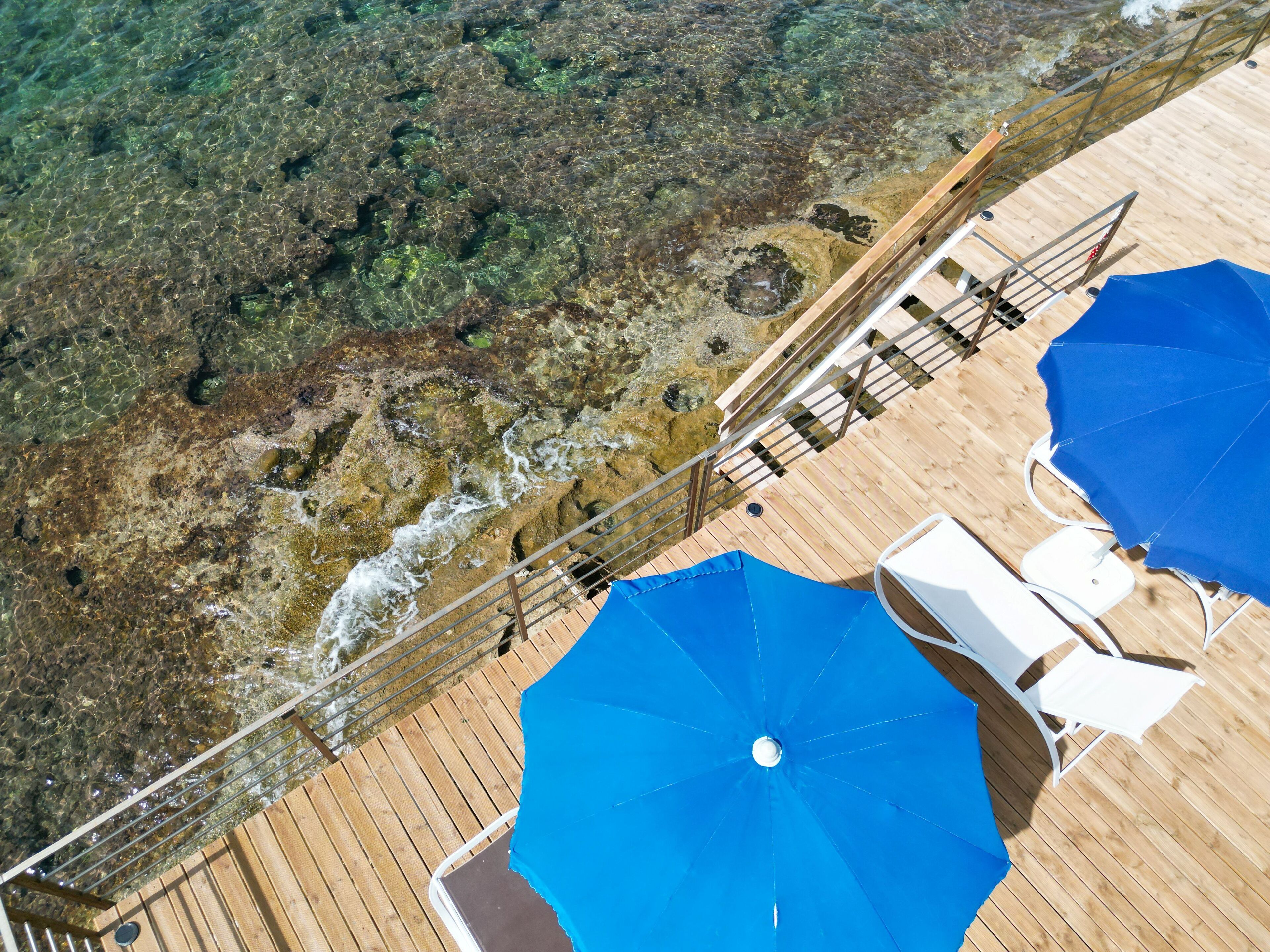 Eine Holzterrasse mit Geländer, einem blauen Sonnenschirm und weißen Liegestühlen.