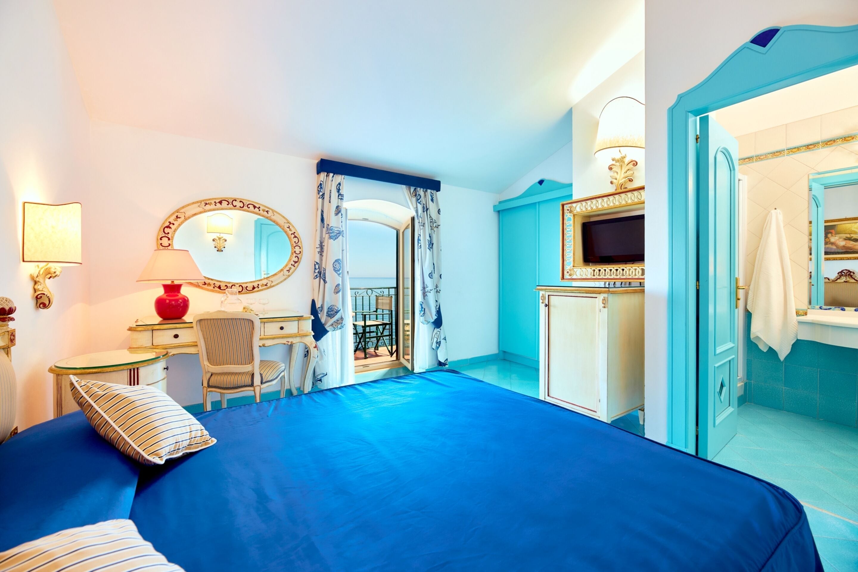 Quarto de hotel com uma cama grande, uma escrivaninha com cadeira, televisão e banheiro com espelho e pia.