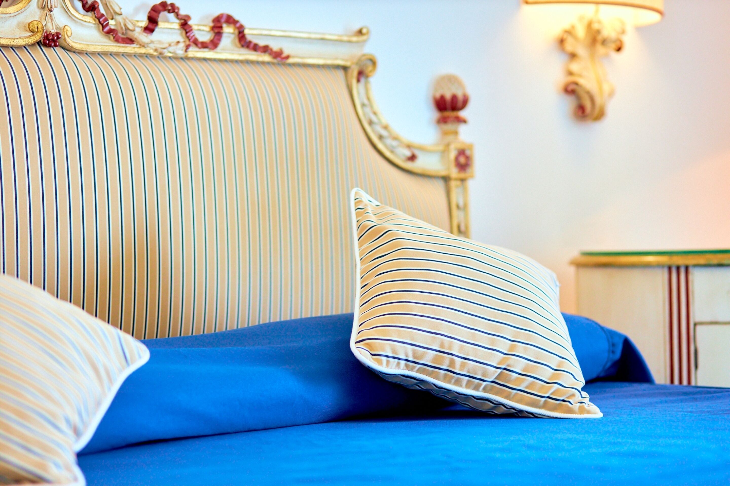 Una cama con ropa de cama azul y almohadas a rayas, un cabecero con marco dorado y una lámpara fijada a la pared.