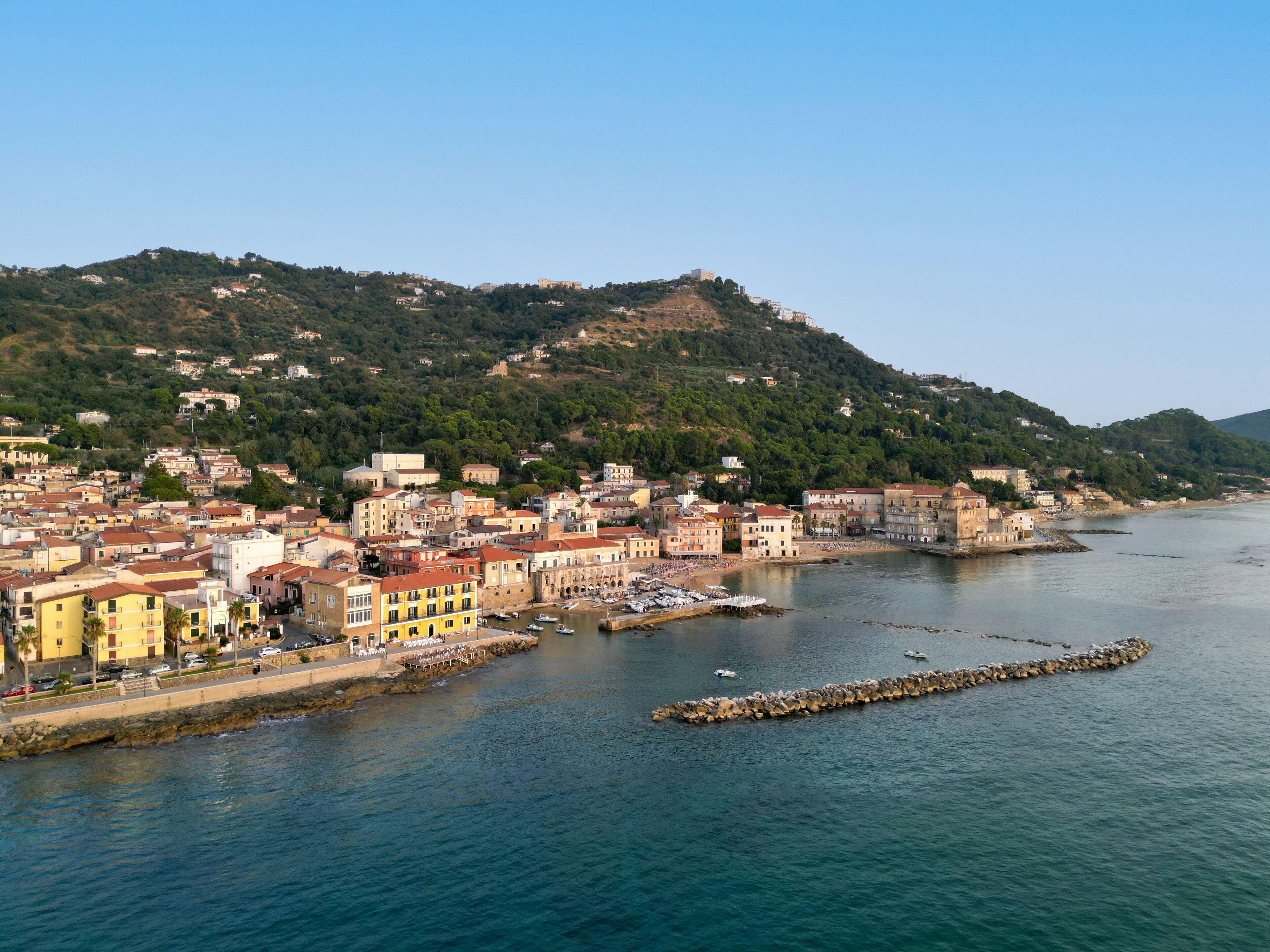 A coastal town with buildings along the waterfront, a rocky breakwater, and a hill with structures in the background.
