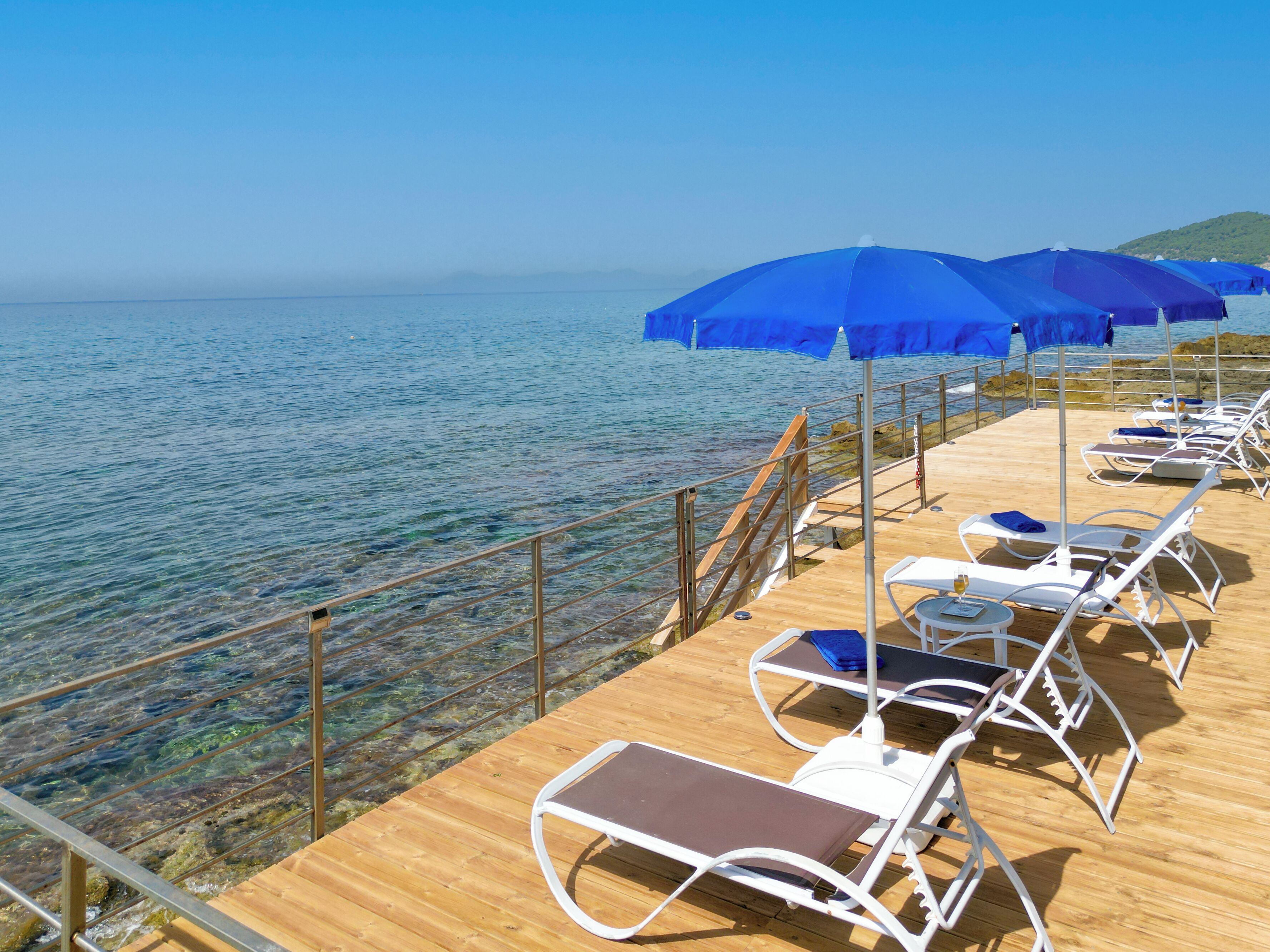 A wooden deck with white lounge chairs and blue umbrellas overlooking clear blue water.