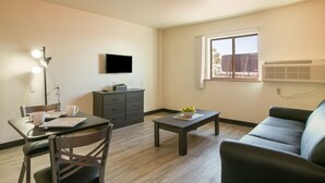 A living room with a sofa, a coffee table, a TV mounted on the wall, and a dining area with chairs and a table. - Siegel Select Albuquerque (Albuquerque)
