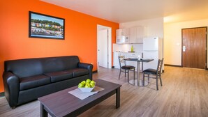 A living room with a black sofa, a coffee table with apples, a dining area with a table and chairs, and a kitchen with white cabinets and appliances. - Siegel Select Albuquerque (Albuquerque)