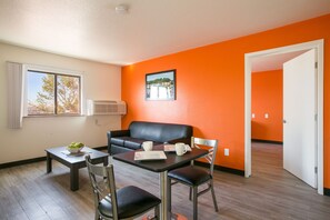 A room with a dining table, chairs, a black leather sofa, a window with blinds, and an air conditioning unit.