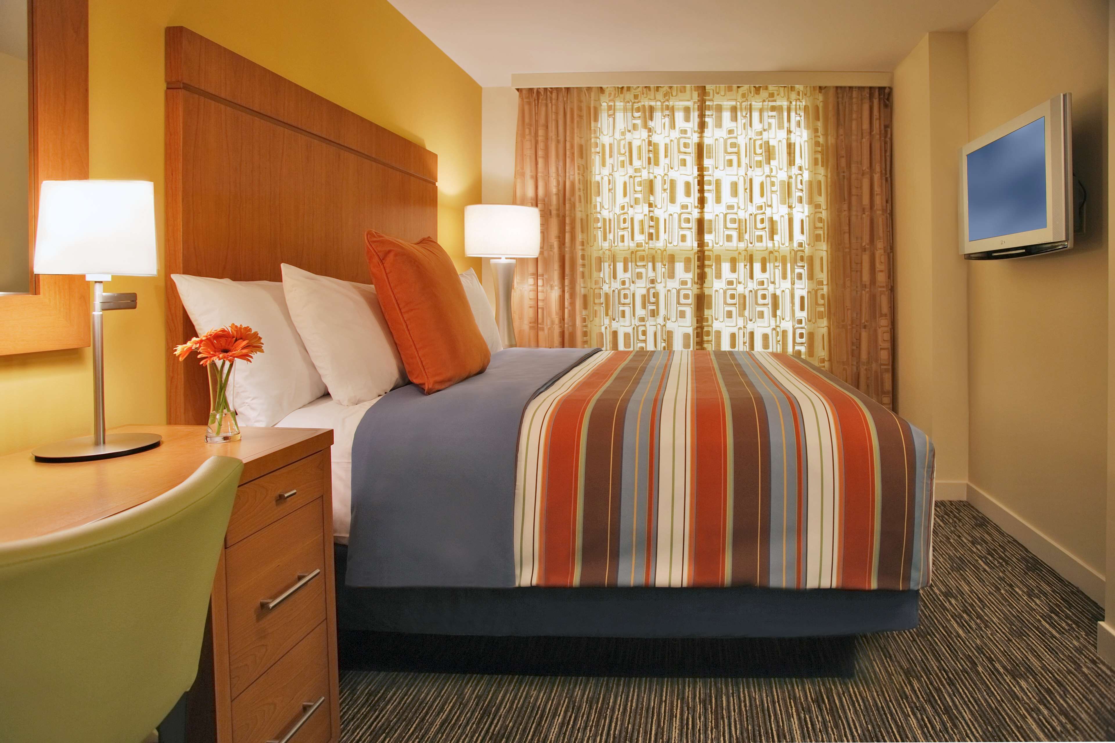 A hotel room with a bed, two bedside lamps, a TV, a desk with a chair, and a vase with flowers.