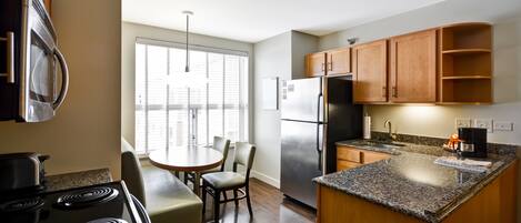 A modern kitchen with a stove, refrigerator, wooden cabinets, granite countertop, and a dining area with chairs and a table.