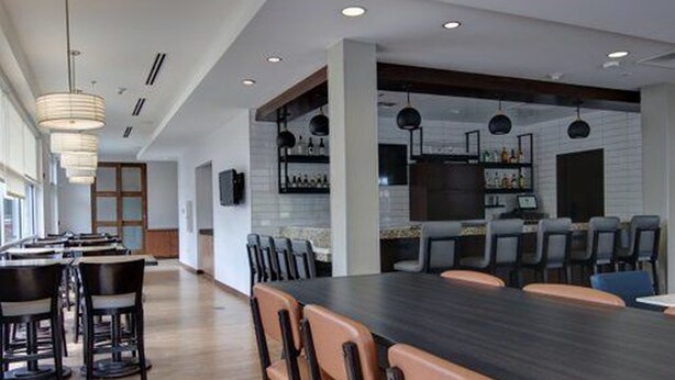 A modern dining area with a long table, multiple chairs, pendant lighting, and a kitchen area in the background. - HYATT house Chicago/Naperville/Warrenville (Warrenville)