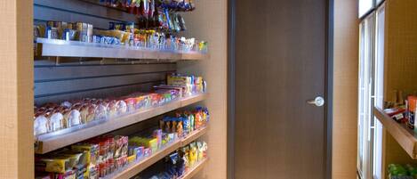 A narrow hallway with shelves stocked with snacks and beverages, a wooden door, and a tiled floor.