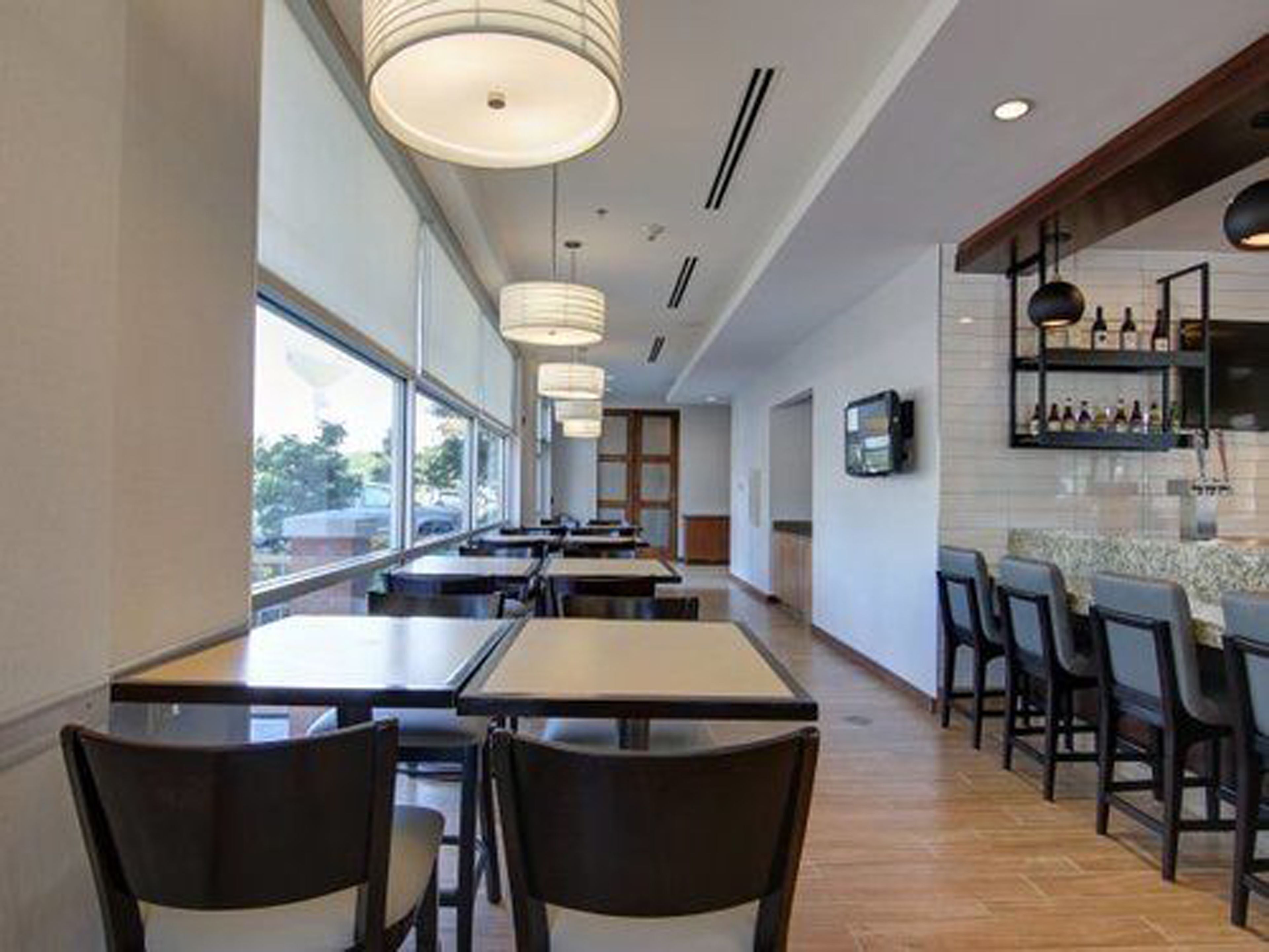 Un interior de restaurante moderno con mesas y sillas largas, iluminación colgante, una zona de cocina con estantería para vinos y grandes ventanales que ofrecen vistas.