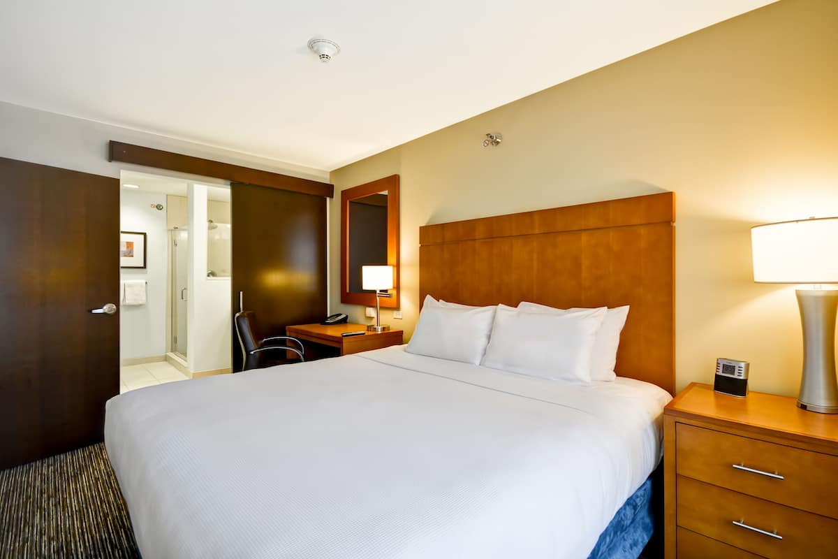 Une chambre d’hôtel avec un grand lit, une table de chevet avec une lampe, un bureau avec une chaise et une salle de bain visible à travers une porte ouverte.