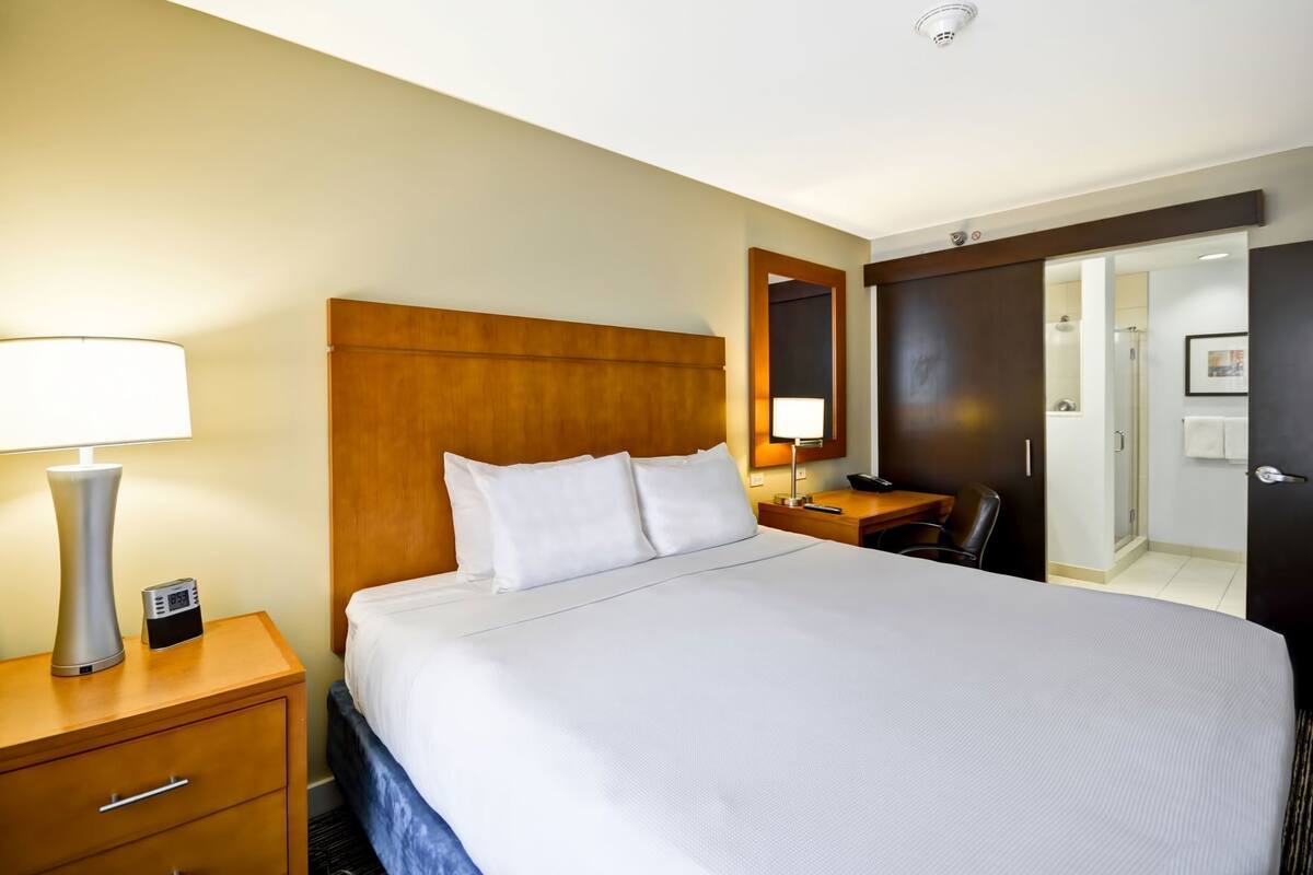 A hotel room with a bed, bedside table, lamp, alarm clock, and a mirror.