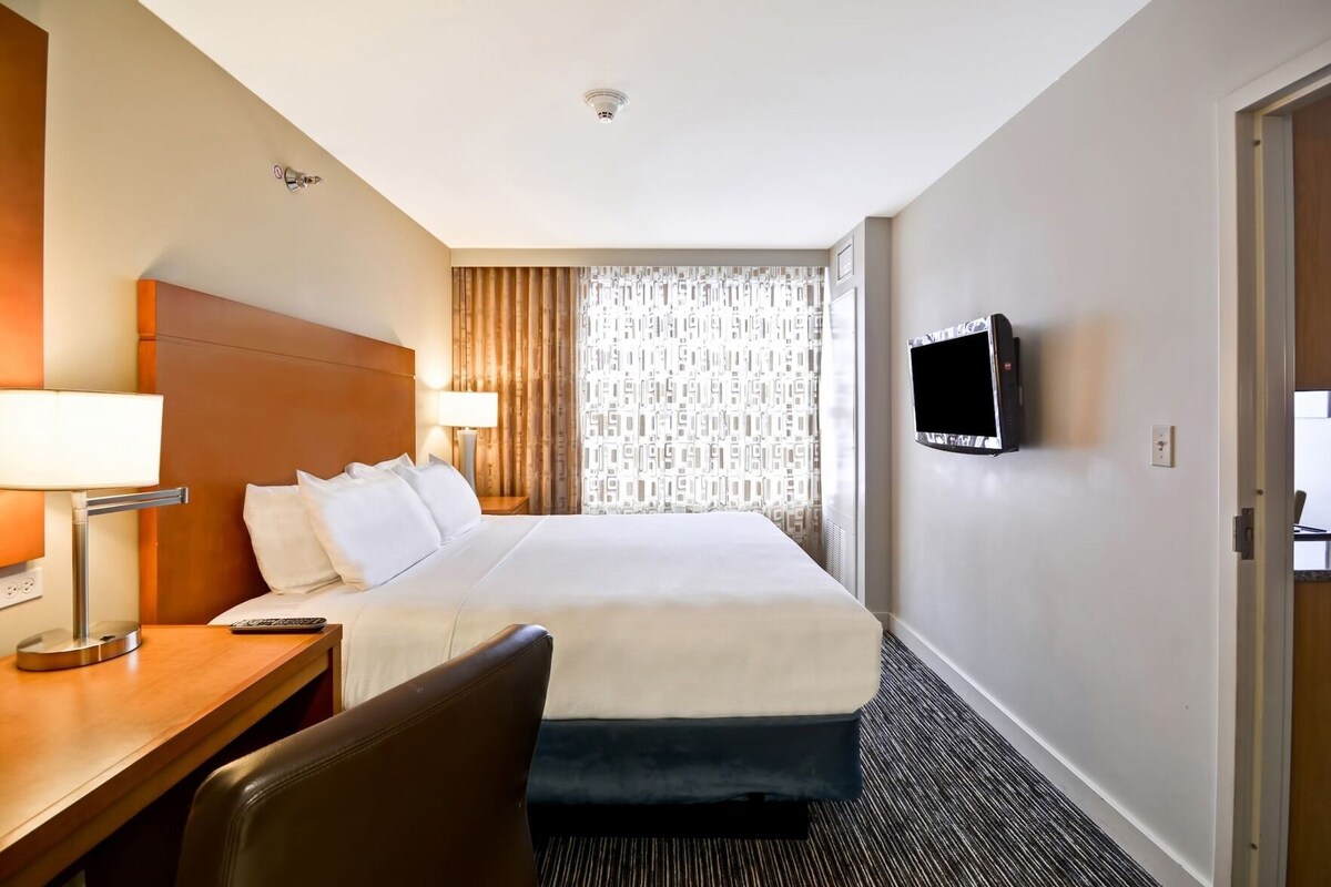 A hotel room with a bed, a desk with a lamp, a chair, a TV mounted on the wall, and a window with curtains.