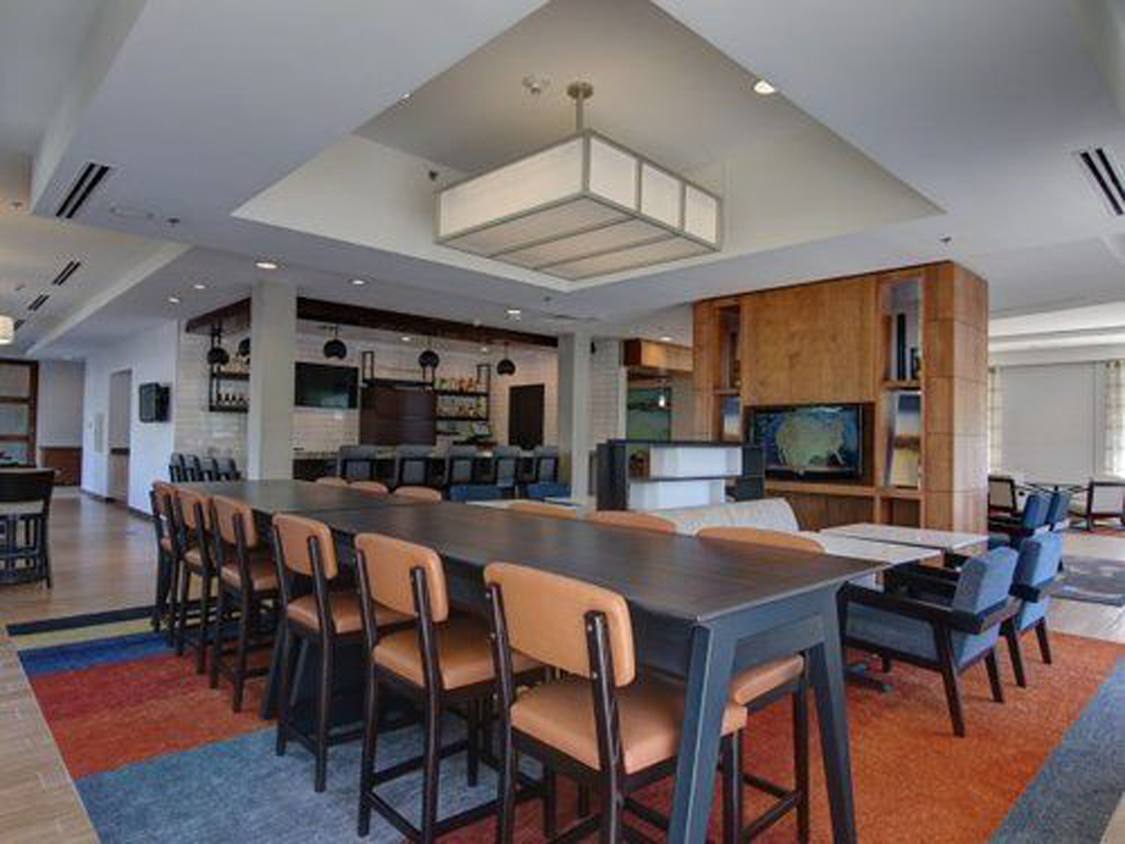 Un espace repas moderne avec une longue table et de nombreuses chaises, un coin cuisine doté d’un grand îlot central, et un coin salon avec un téléviseur et des étagères à livres.