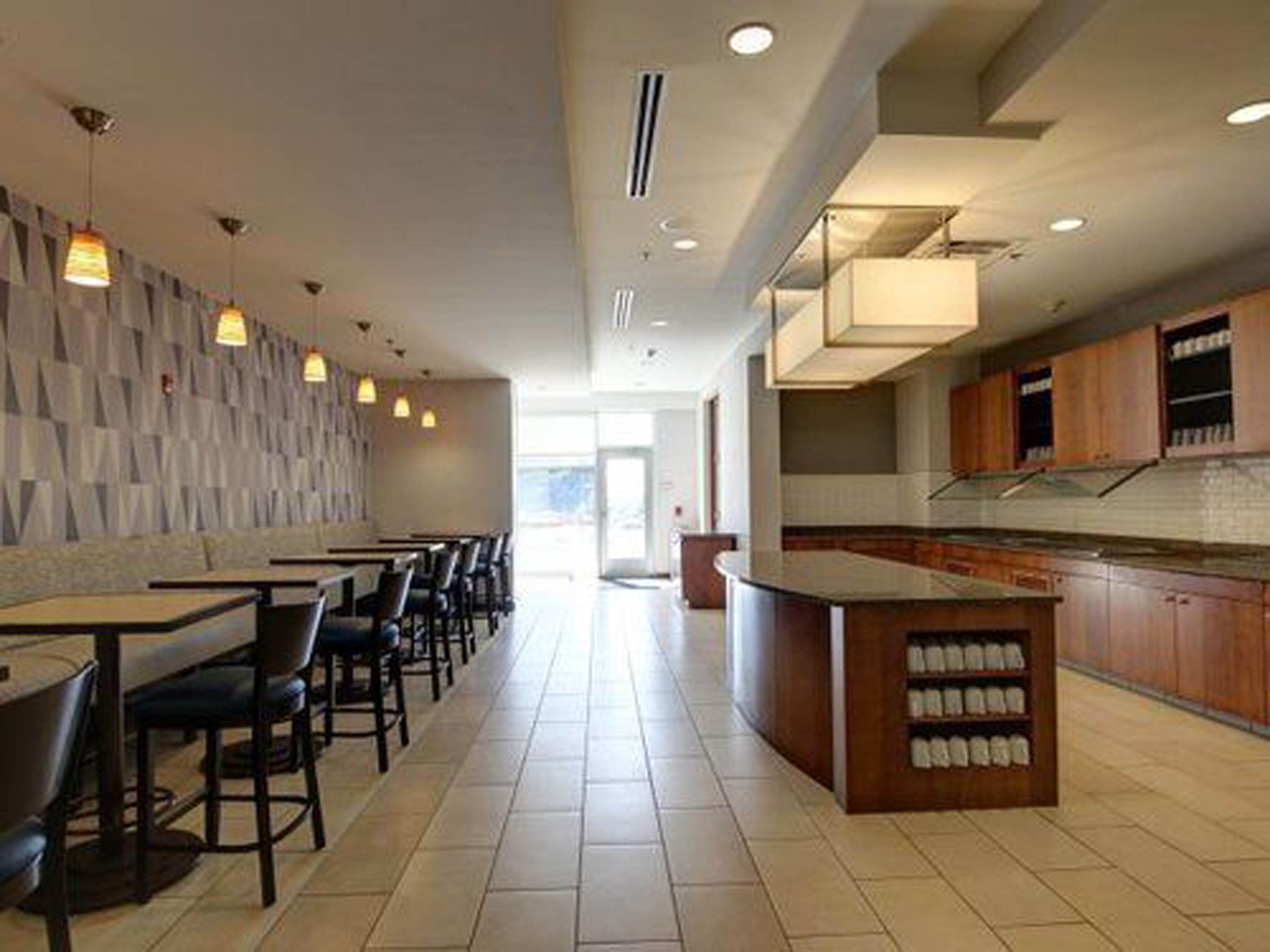 Un restaurante moderno con una barra alargada, varias mesas y sillas. Se observa una zona de cocina con armarios de madera y una exhibición de botellas.