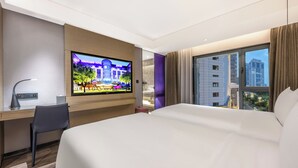 A hotel room with a large bed, a desk with a chair, a television displaying a hotel exterior, and a view of city buildings through the window. - Mercure Harbin Central Street Sophia Church (Harbin)