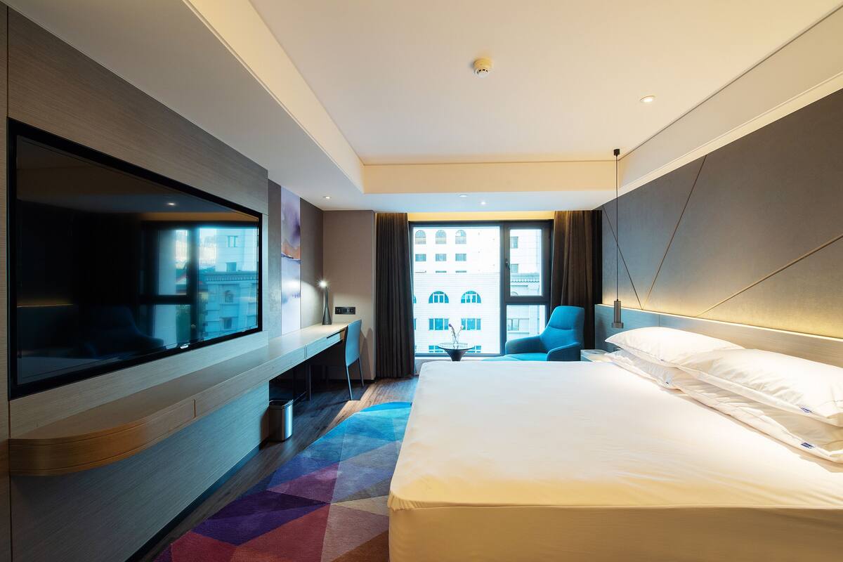 A modern hotel room with a large bed, a flat-screen TV, a desk, and a view of the city through the window.